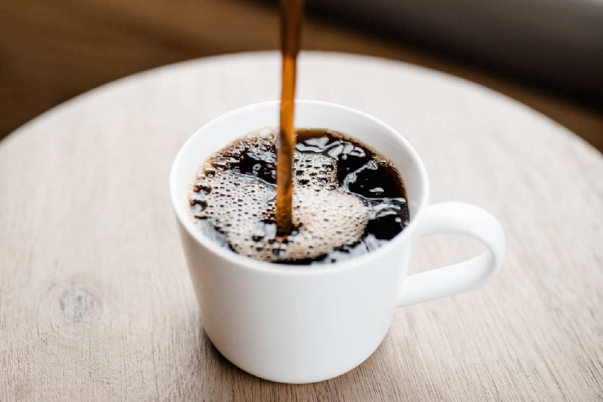 Una tazzina da caffè è sopra ad un tavolino di legno e dall'alto viene versata la bevanda scura fino proprio dentro la tazza